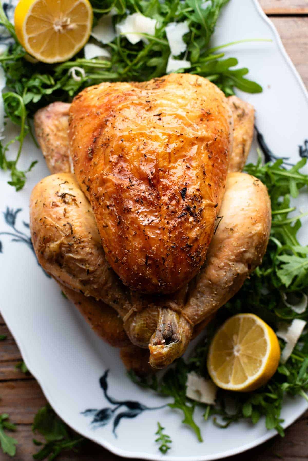 A close up of a roast chicken on a serving plate with arugula and lemons.