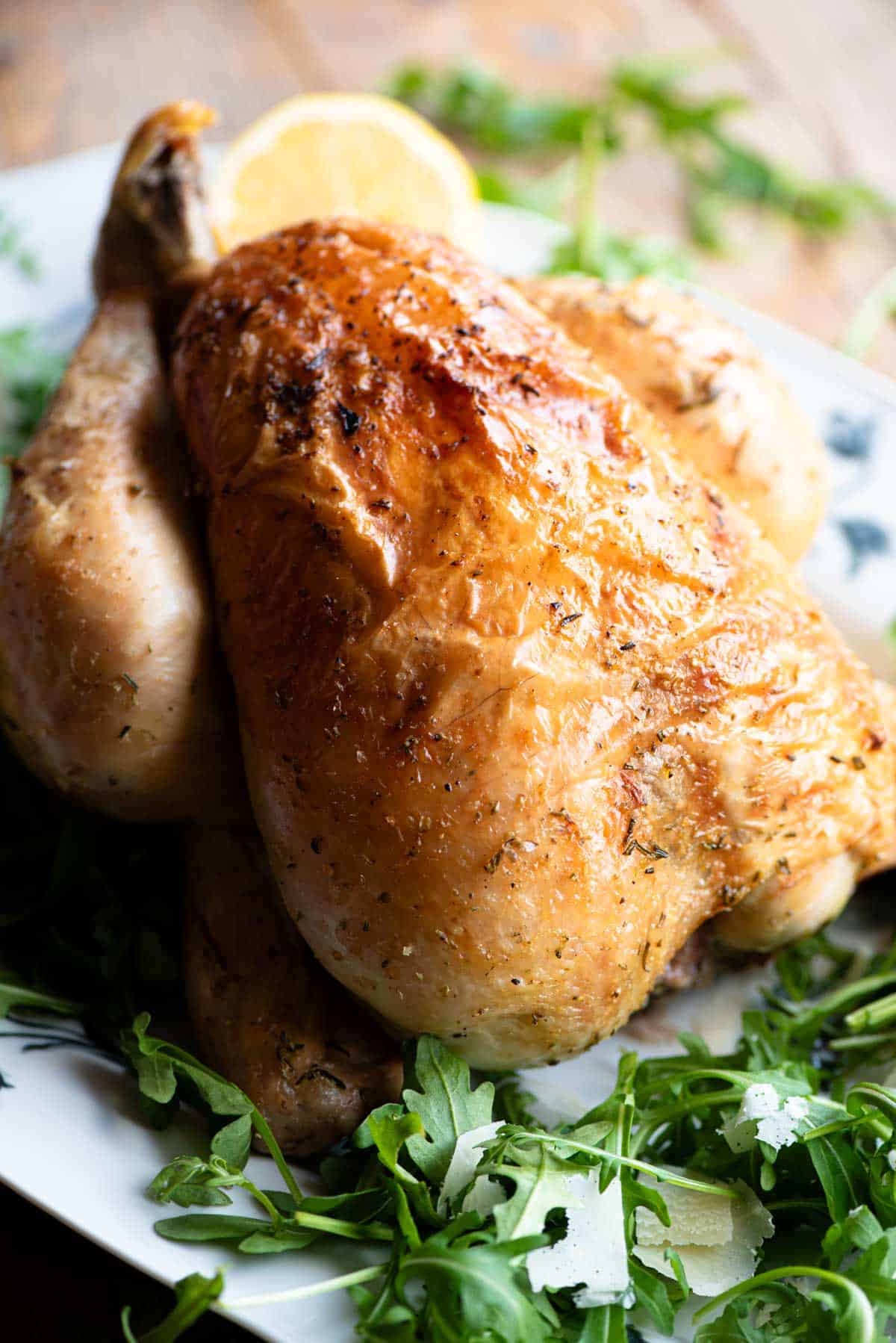 A close up side shot of a whole lemon roast chicken on a serving plate with arugula and lemon slices around it.