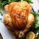 A close up cropped image of a roast chicken on a plate with lemon and arugula.