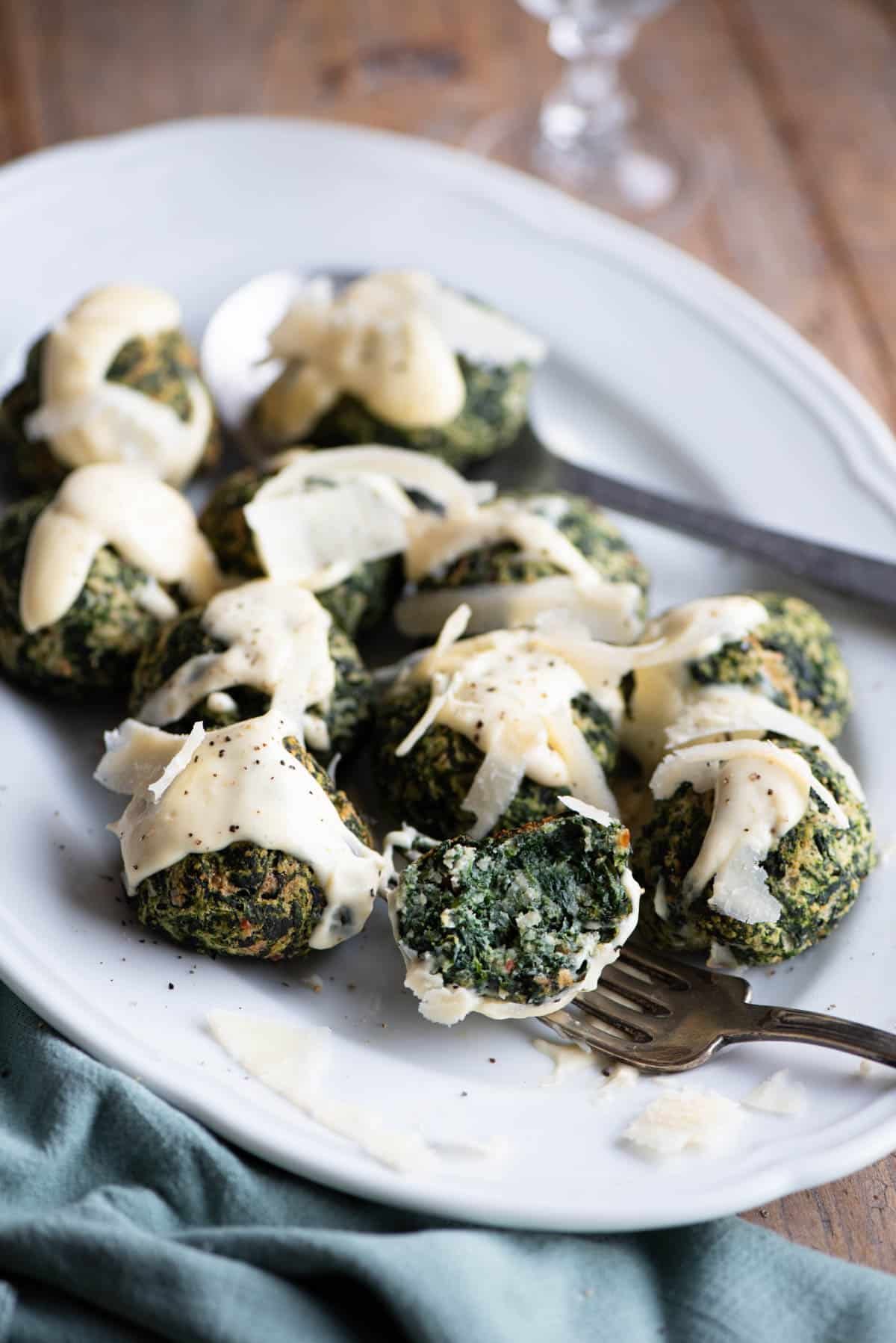 Spinach balls on a white serving plate with parmesan cream. One is cut open on a fork.
