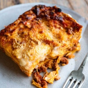 A close up of a slice of lamb lasagna on a plate with a fork at the side.