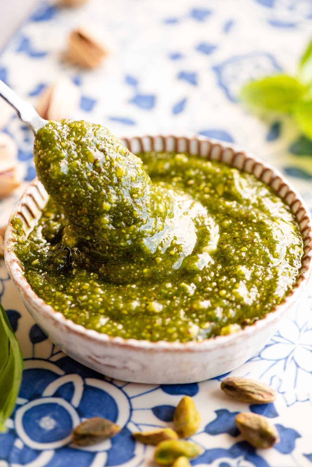 A close up of a spoon in a dish of pistachio pesto sitting on a blue patterned background with pistachios around.