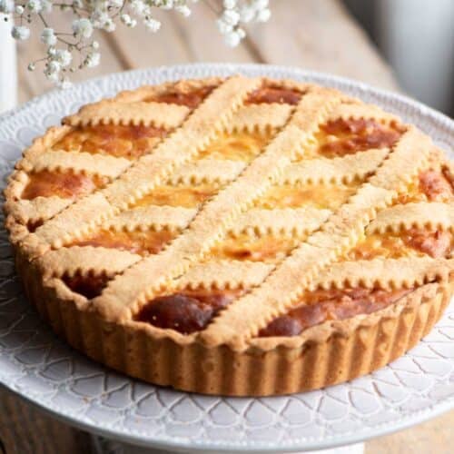 Pastiera Napoletana on a cake stand sitting on a wooden surface with white flowers at the side.
