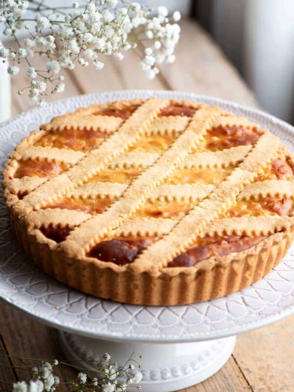 Pastiera Napoletana on a cake stand sitting on a wooden surface with white flowers at the side.