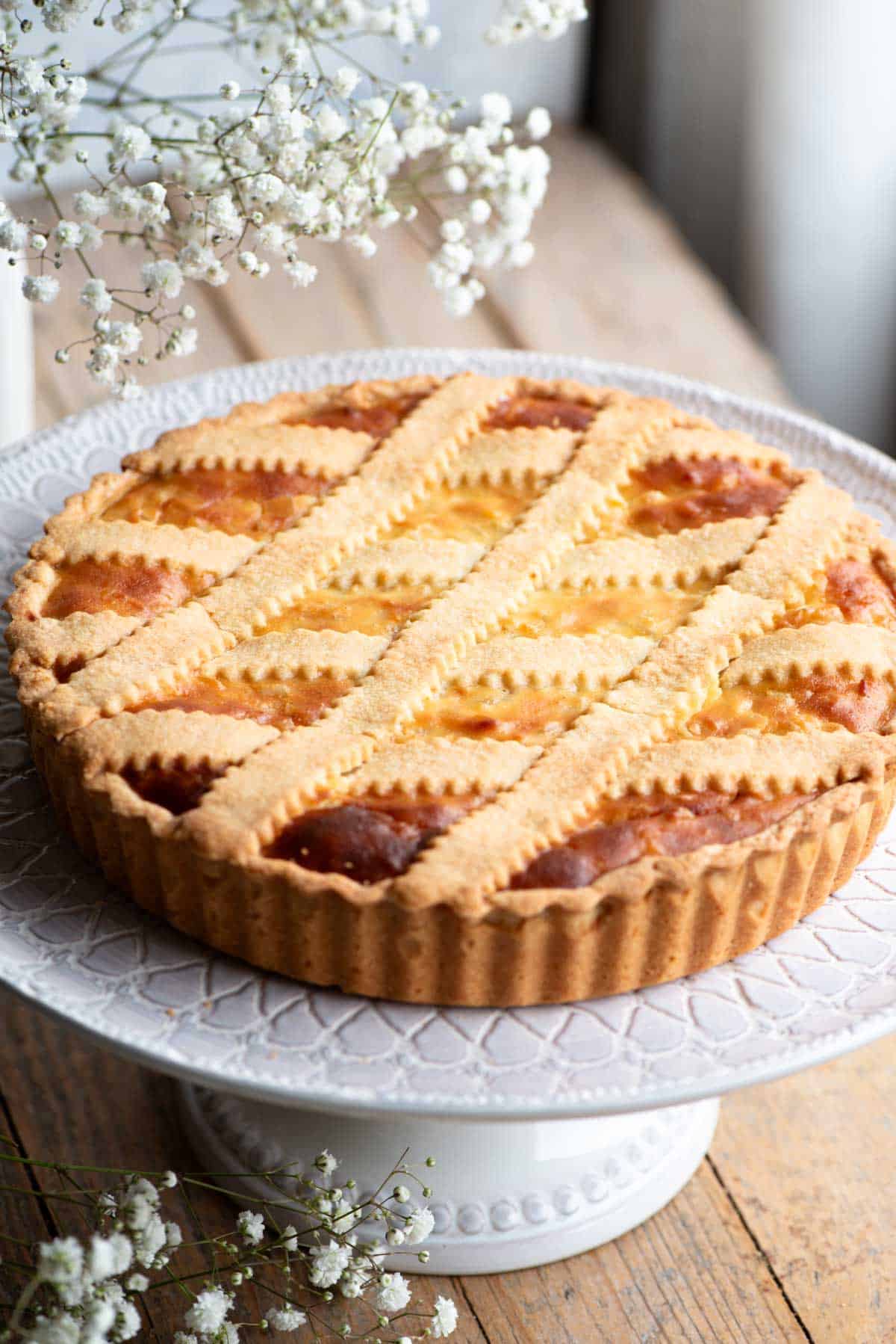 Pastiera Napoletana on a cake stand sitting on a wooden surface with white flowers at the side.