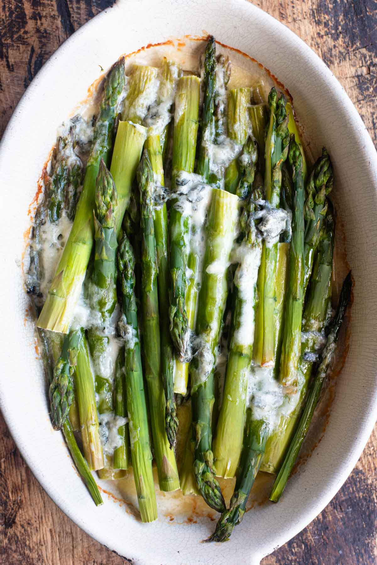 An overhead shot of roasted asparagus in an oval baking dish topped with melted gorgonzola cheese.