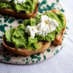 A close up of a slice of bruschetta topped with pea pesto and goats cheese on a ceramic serving board.