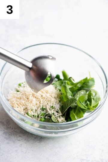 All ingredients for pea pesto in a bowl with an immersion blender over it.