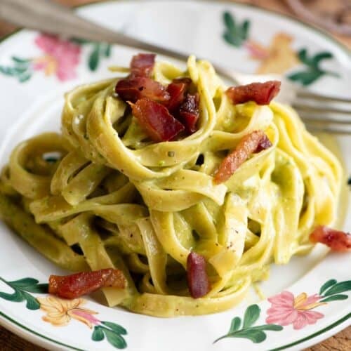 Creamy pistachio pasta on a floral patterned plate topped with guanciale. The background is wooden.