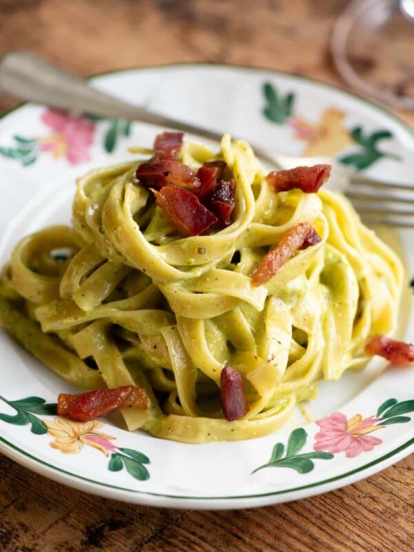 Creamy pistachio pasta on a floral patterned plate topped with guanciale. The background is wooden.