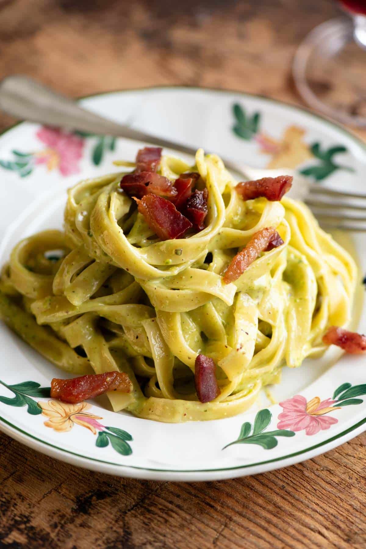 Creamy pistachio pasta on a floral patterned plate topped with guanciale. The background is wooden.