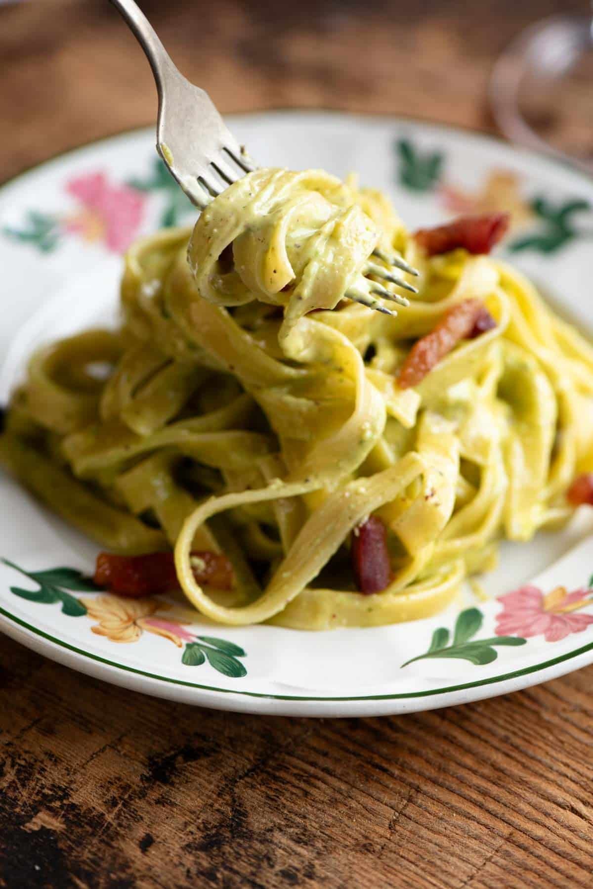 A fork picking up some tagliatelle with pistachio cream sauce from a floral patterned plate.