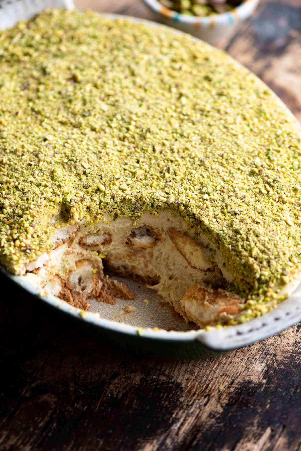 A side shot of a pistachio tiramisu in an oval dish with a slice removed. The background is wooden.