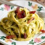 A cropped square image of pistachio pasta on a floral patterned plate topped with crispy guanciale (bacon).