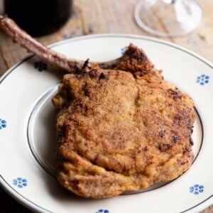 A breaded veal chop sitting on a plate with a glass and bottle of red wine in the background.
