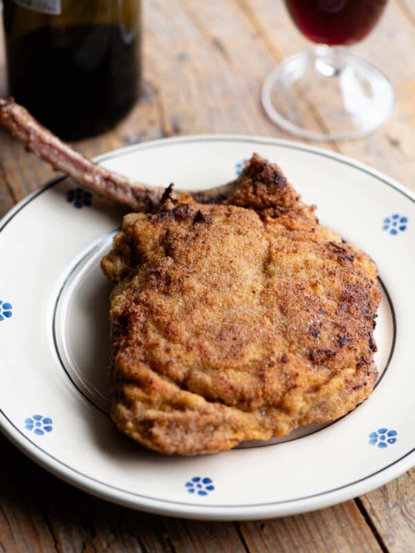 A breaded veal chop sitting on a plate with a glass and bottle of red wine in the background.