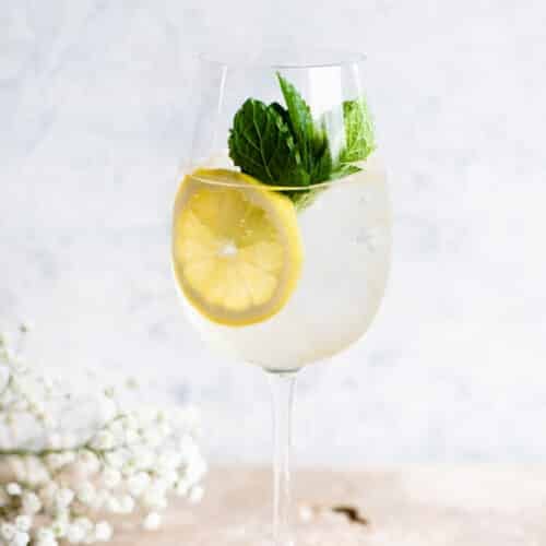 A Hugo spritz cocktail on a light coloured worktop with lemon and mint.