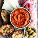 A cropped image of Ajvar, a red pepper dip in a blue bowl surrounded by antipasti.