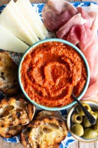 A close up of Ajvar dip in a blue bowl surrounded by cured meat, cheese, ciabatta bread and olives.