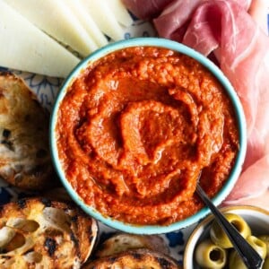 A close up of Ajvar dip in a blue bowl surrounded by cured meat, cheese, ciabatta bread and olives.
