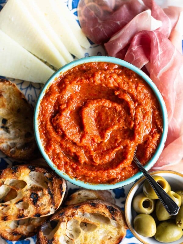A close up of Ajvar dip in a blue bowl surrounded by cured meat, cheese, ciabatta bread and olives.