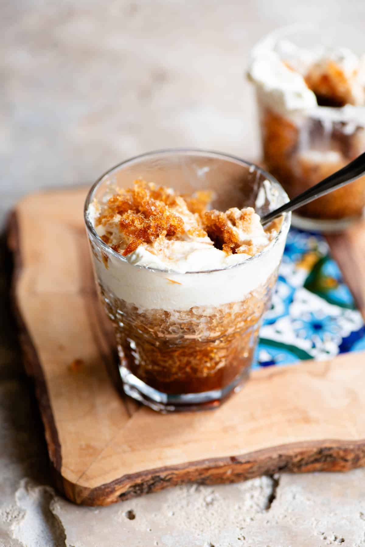 A glass filled with coffee granita and whipped cream sitting on a wooden serving board with a patterned tile in the middle.