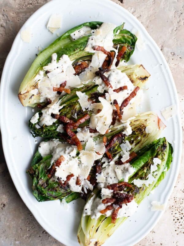An overhead shot of a white serving plate with grilled romaine lettuce topped with a creamy sauce, pancetta and Parmigiano Reggiano shavings.