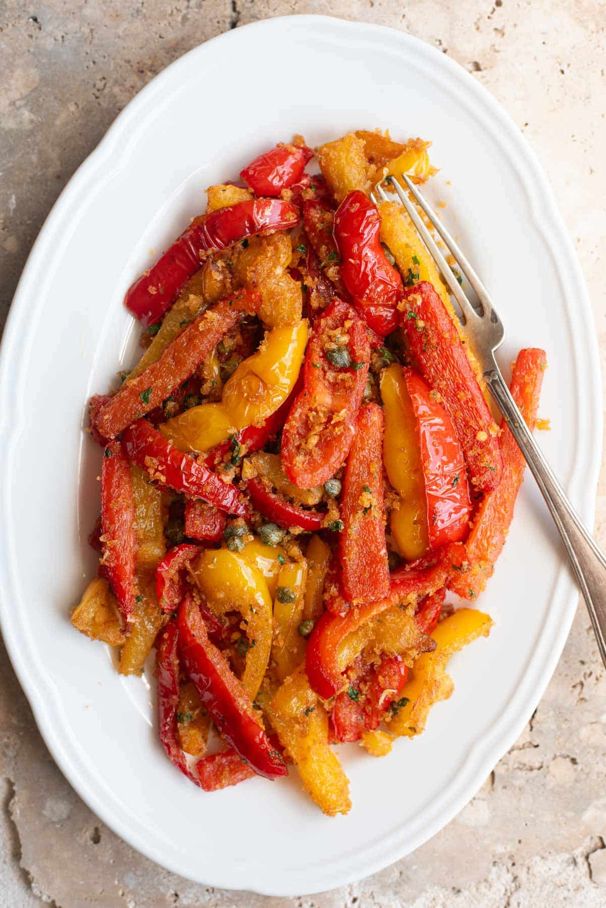 An overhead shot of peppers, capers and breadcrumbs on a white oval serving dish.