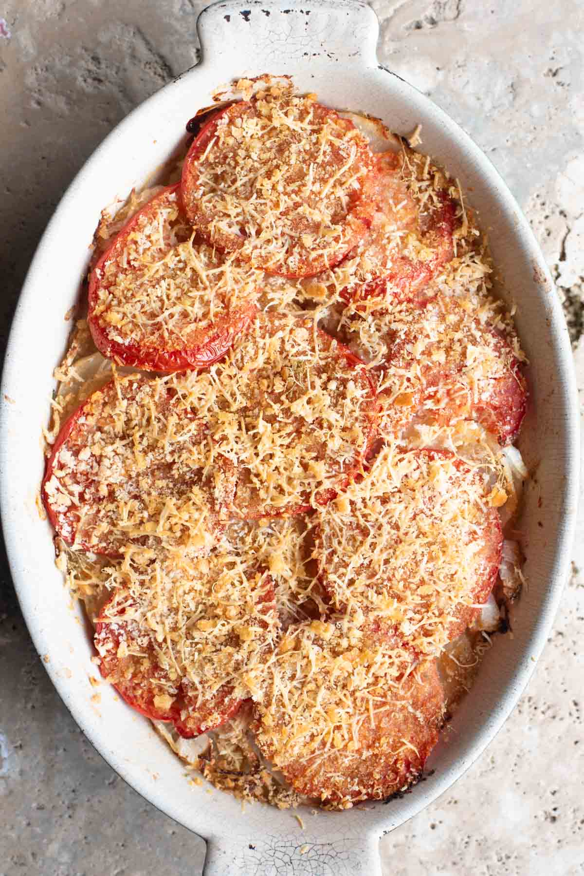 An overhead shot of an Italian potato and tomato gratin in an oval dish sitting on a Travertine background.
