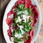 A close up cropped image of beef carpaccio on a white serving plate with arugula and shavings of Grana Padano cheese.