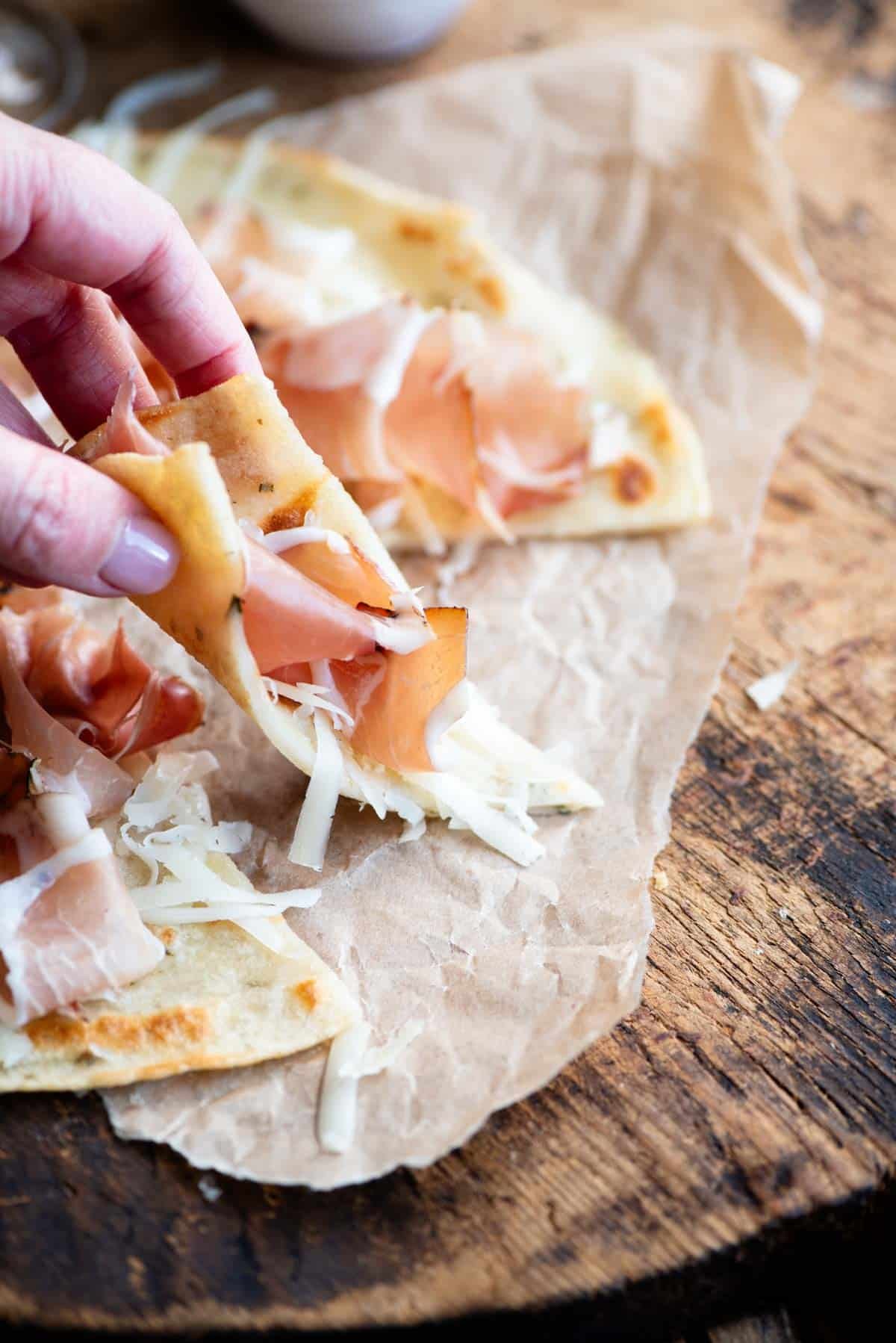 A hand picking up a slice of Italian flatbread topped with cured meat and cheese.