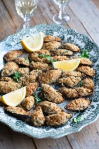Italian stuffed mussels on a large serving plate with lemon wedges and 2 glasses of wine in the background.