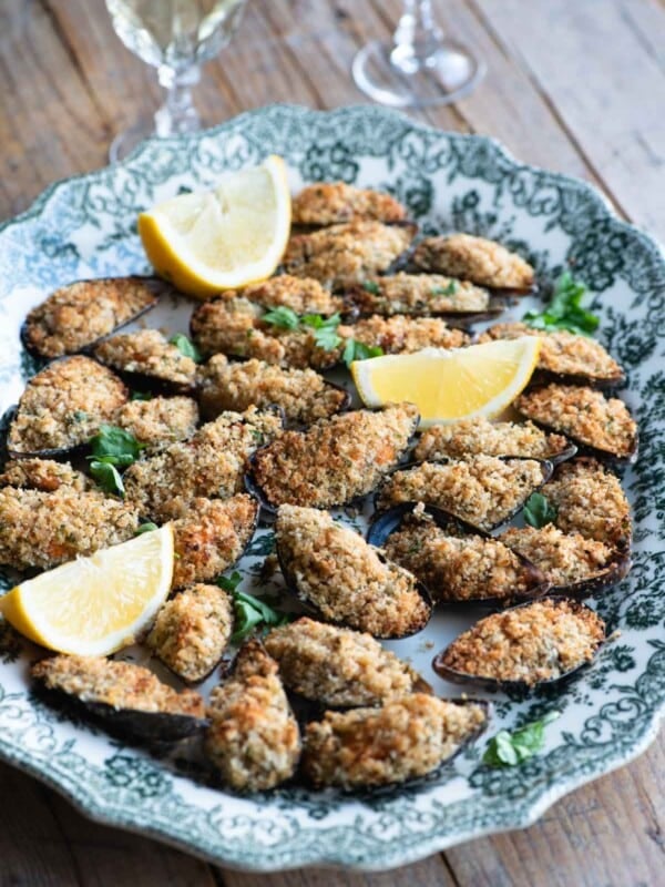 Italian stuffed mussels on a large serving plate with lemon wedges and 2 glasses of wine in the background.