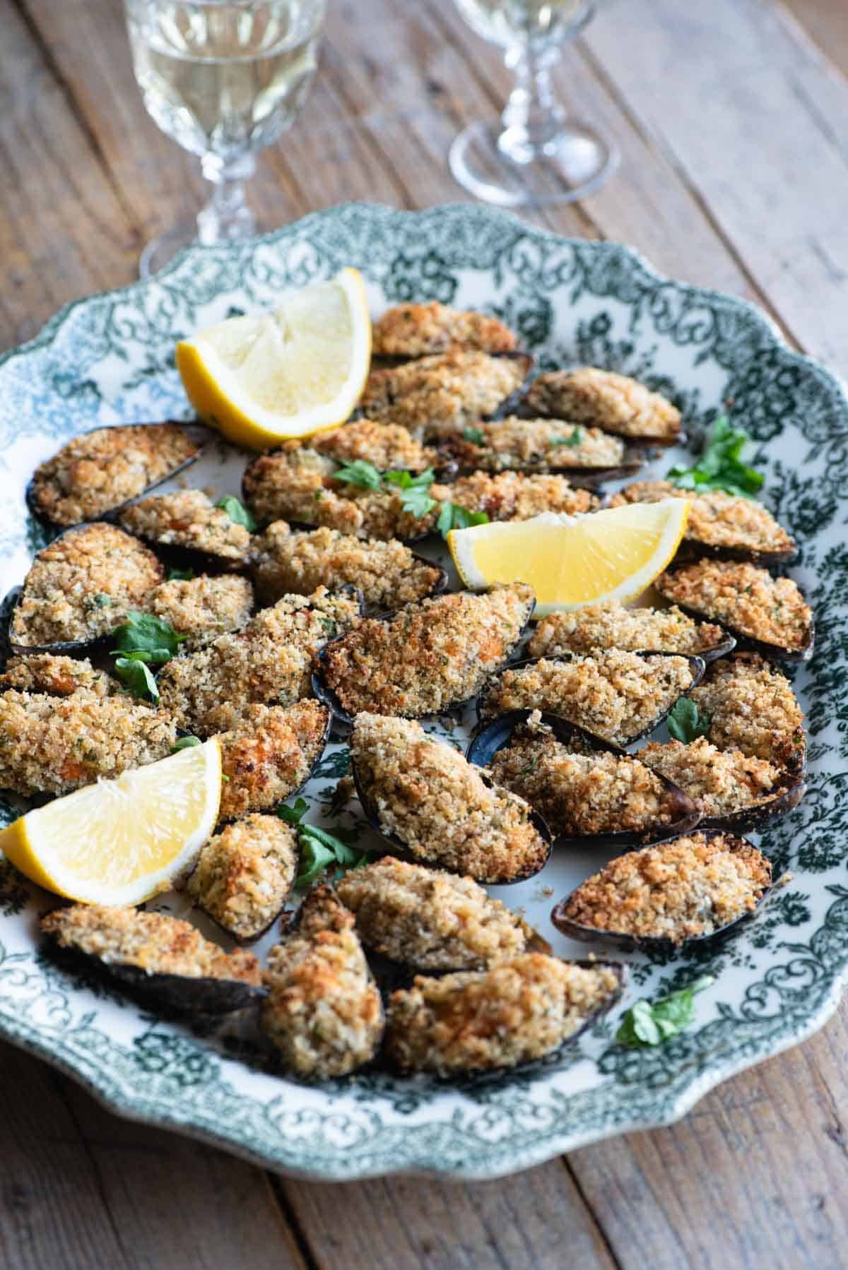 Italian stuffed mussels on a large serving plate with lemon wedges and 2 glasses of wine in the background.