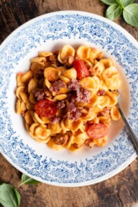 An overhead shot of pasta with sausage, tomato and burrata in a blue and white bowl.