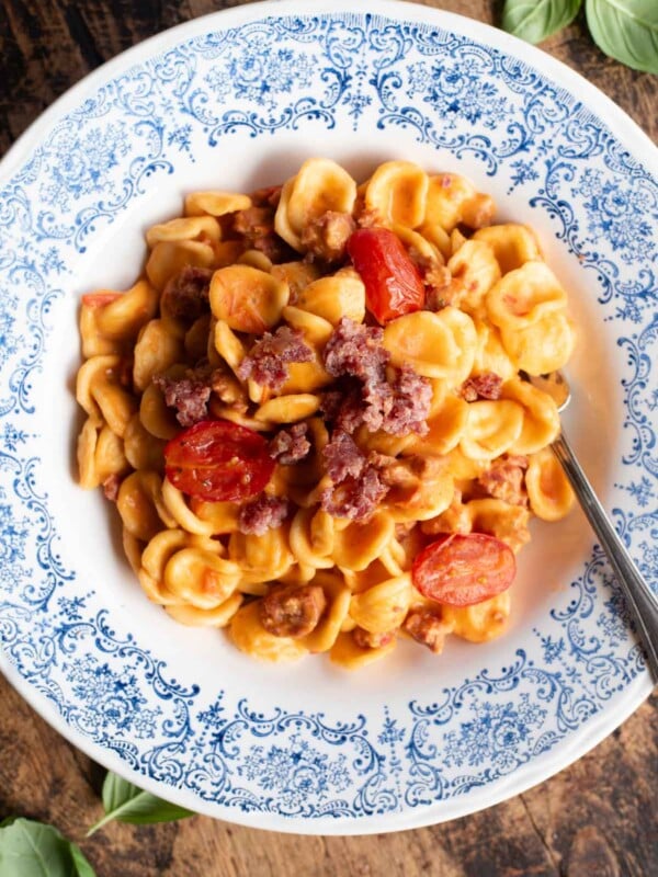 An overhead shot of pasta with sausage, tomato and burrata in a blue and white bowl.