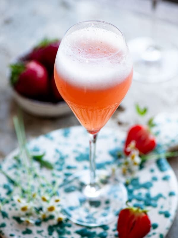 A close up of a strawberry bellini (rossini) cocktail sitting on travertine with strawberries and flowers around.