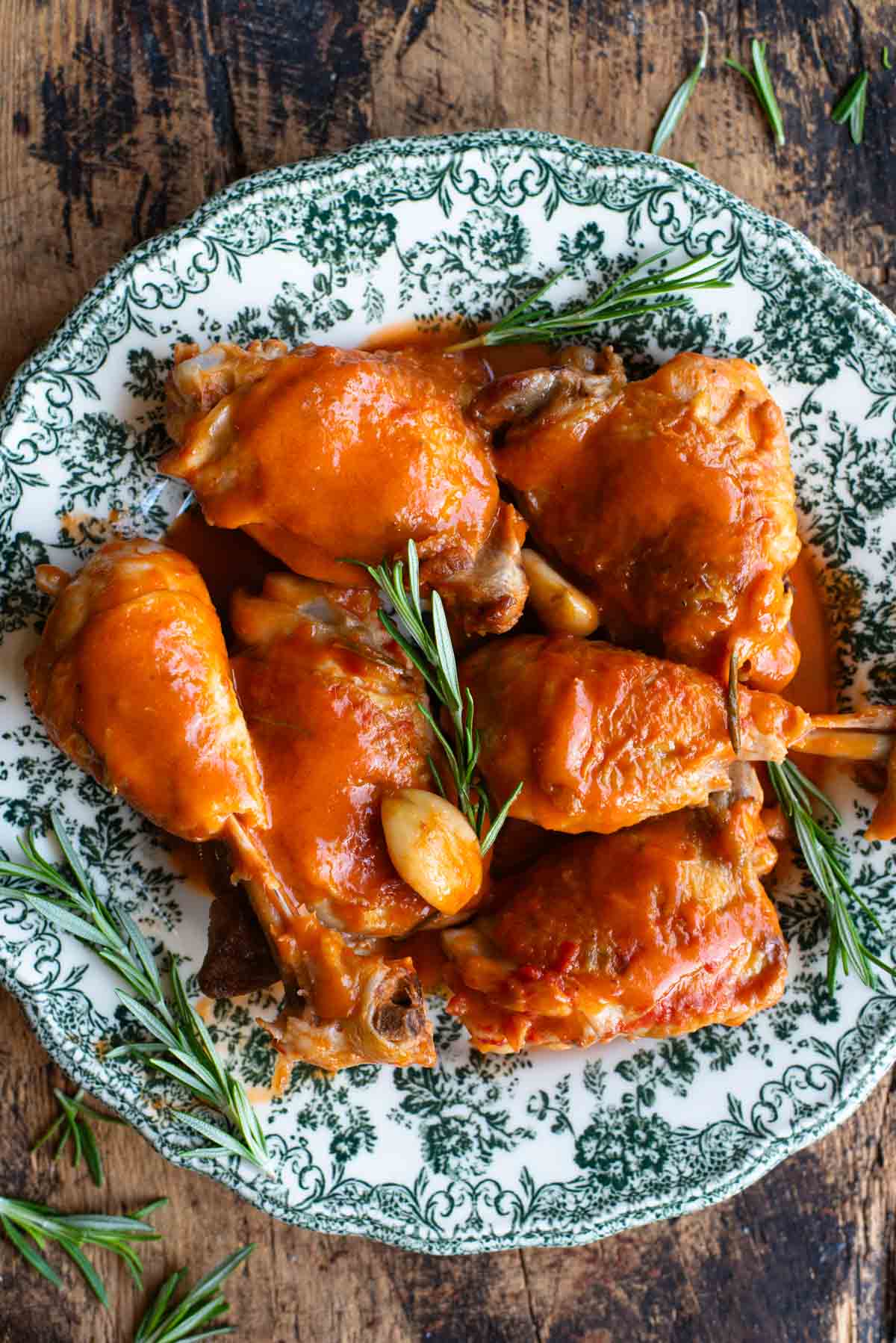 An overhead shot of chicken thighs and drumsticks on a green patterned plate with rosemary and garlic.