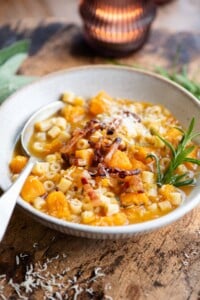 A butternut squash and pasta soup topped with pancetta and a sprig of rosemary. The bowl is on a wooden table.