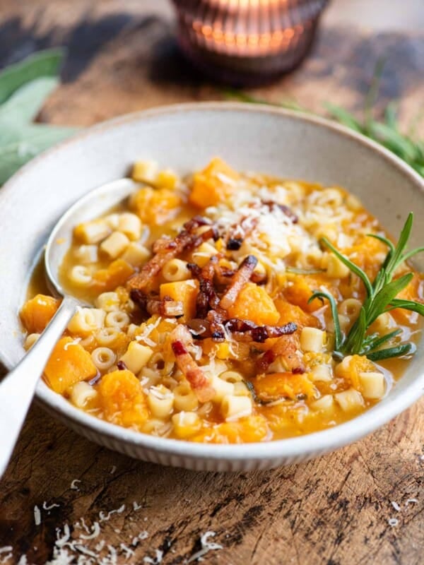 A butternut squash and pasta soup topped with pancetta and a sprig of rosemary. The bowl is on a wooden table.