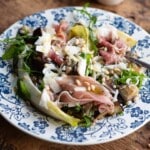 A close up cropped image of an eggplant and farro salad on a blue plate with prosciutto and pine nuts.