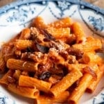 A close up of a bowl of pasta with tomato sauce, guanciale and pecorino on a wooden surface.
