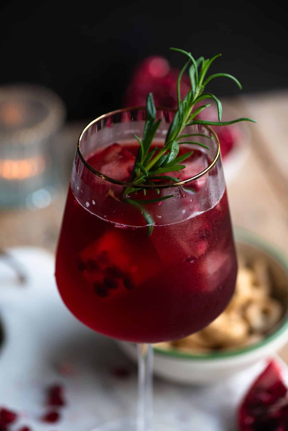 A close up of a pomegranate spritz in a wine glass with a rosemary sprig.