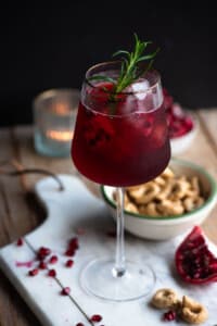 A pomegranate spritz in a tall wine glass with a gold rim and a sprig of rosemary inside.
