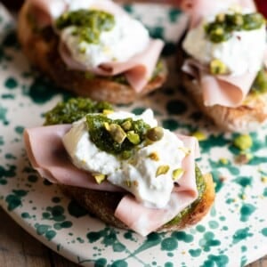 A close up of three crostini on a spotted serving board topped with mortadella, burrata and pistachios.