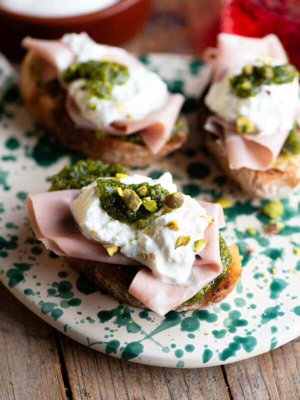 A close up of three crostini on a spotted serving board topped with mortadella, burrata and pistachios.