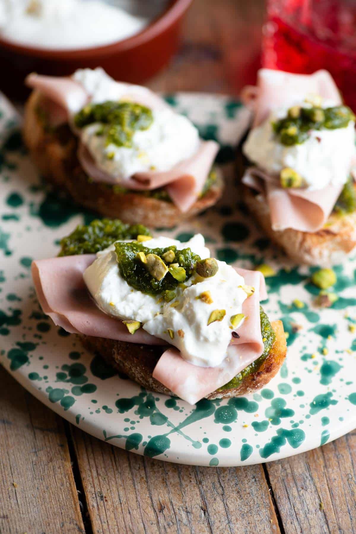 A close up of three crostini on a spotted serving board topped with mortadella, burrata and pistachios.