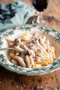 Pasta alla Norcina in a green and white bowl with a fork and glass of wine in the background.