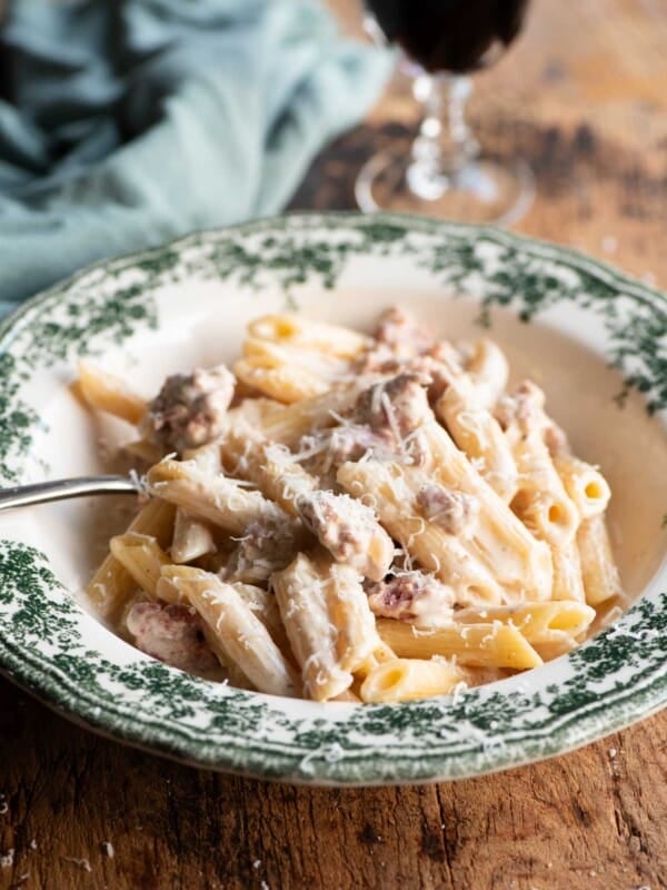 Pasta alla Norcina in a green and white bowl with a fork and glass of wine in the background.