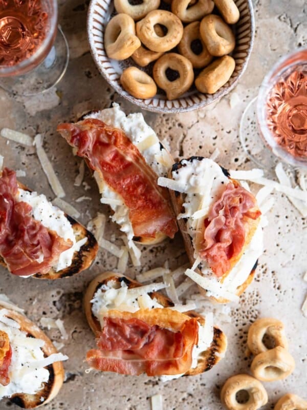 An overhead shot of burrata and Parmigiano crostini on a travertine background topped with prosciutto.